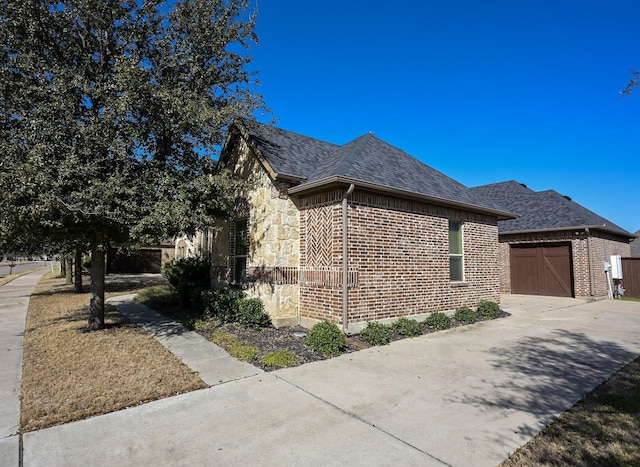 view of side of property featuring driveway, stone siding, a shingled roof, a garage, and brick siding