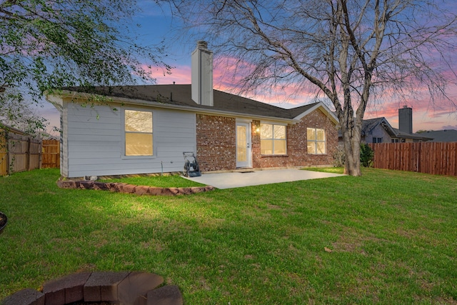 back of property at dusk featuring a fenced backyard, a chimney, a lawn, a patio area, and brick siding