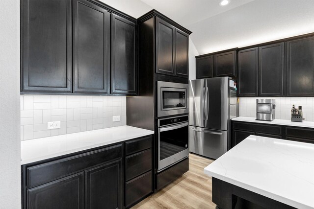 kitchen with light stone countertops, light wood-type flooring, decorative backsplash, appliances with stainless steel finishes, and dark cabinets