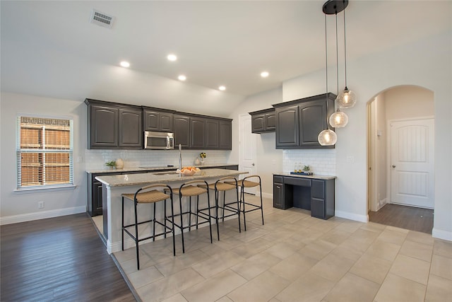 kitchen with visible vents, a center island with sink, stainless steel microwave, arched walkways, and light stone countertops