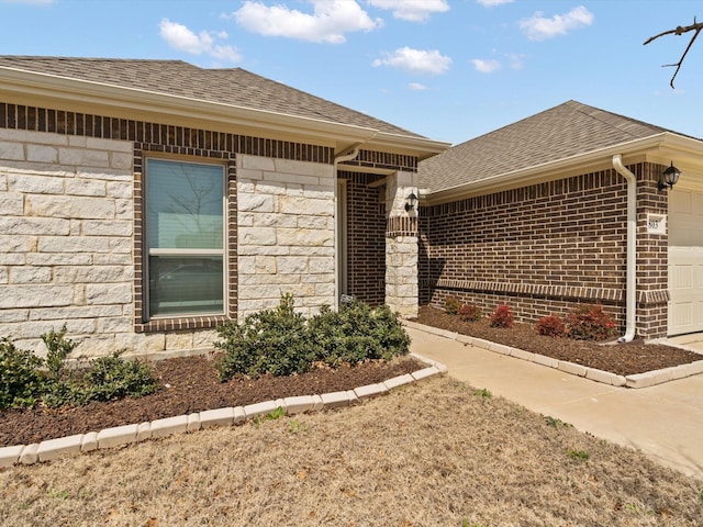 view of exterior entry featuring an attached garage, brick siding, stone siding, and roof with shingles