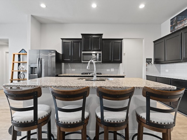 kitchen with light wood-style flooring, an island with sink, a sink, stainless steel appliances, and dark cabinets