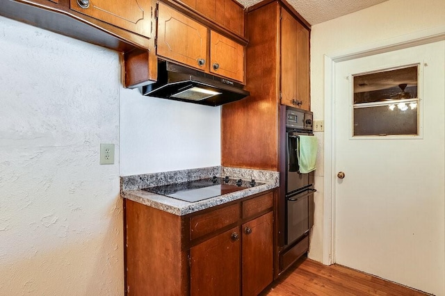 kitchen with under cabinet range hood, brown cabinets, light wood-style floors, and black appliances