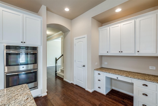 kitchen with arched walkways, stainless steel double oven, white cabinets, built in study area, and dark wood-style flooring