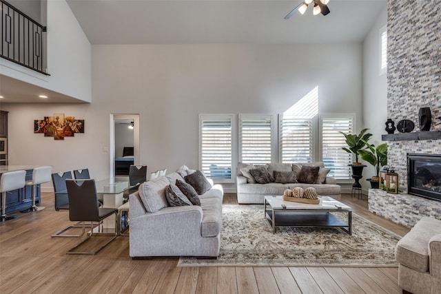 living room with a towering ceiling, a stone fireplace, a ceiling fan, and wood finished floors