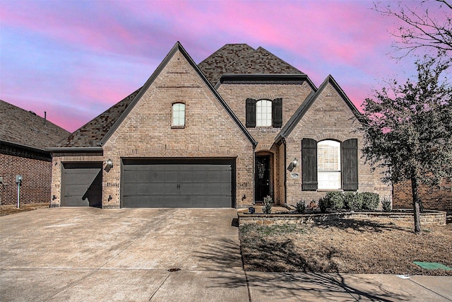 french country inspired facade with brick siding, an attached garage, and concrete driveway