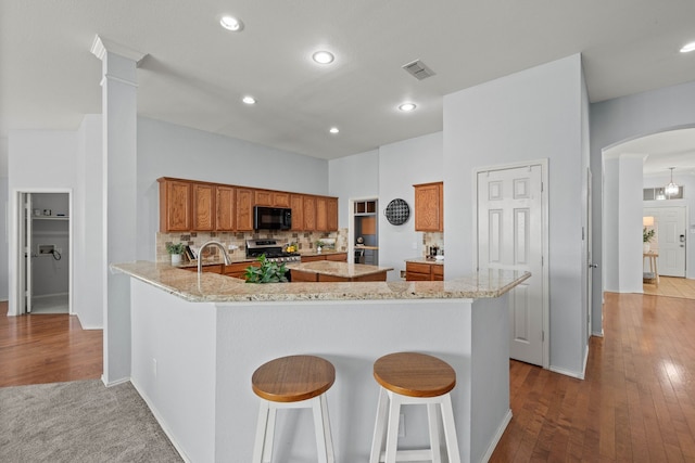 kitchen featuring brown cabinets, arched walkways, black microwave, decorative backsplash, and stainless steel gas range