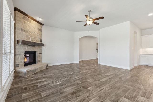 unfurnished living room with baseboards, ceiling fan, a stone fireplace, wood finished floors, and arched walkways