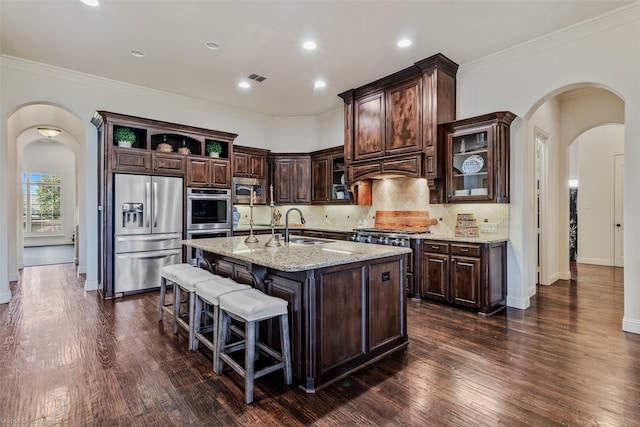 kitchen featuring a kitchen island with sink, open shelves, tasteful backsplash, stainless steel appliances, and arched walkways