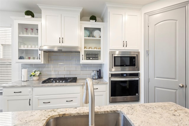 kitchen featuring tasteful backsplash, glass insert cabinets, under cabinet range hood, appliances with stainless steel finishes, and white cabinetry