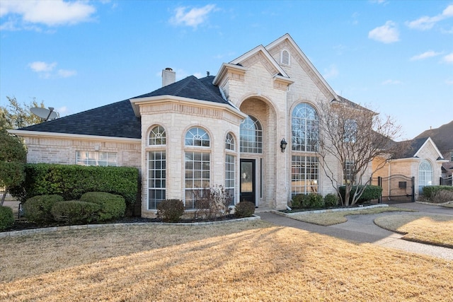 french provincial home with brick siding, a front yard, a chimney, stone siding, and a gate