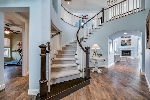 staircase featuring wood finished floors, ceiling fan, a fireplace with raised hearth, and baseboards