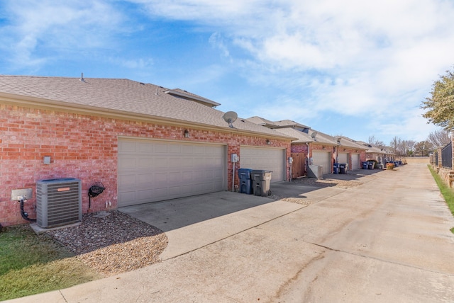 view of side of property with cooling unit, driveway, an attached garage, a shingled roof, and brick siding