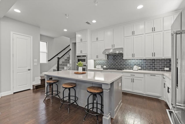 kitchen with stainless steel appliances, dark wood-type flooring, under cabinet range hood, white cabinetry, and tasteful backsplash