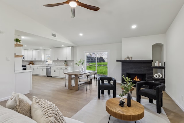 living room with visible vents, light wood-style flooring, a ceiling fan, a brick fireplace, and vaulted ceiling