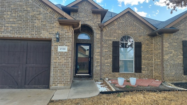 view of exterior entry featuring brick siding, a shingled roof, and a garage