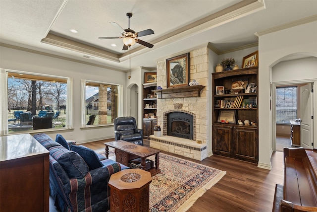 living area with dark wood finished floors, arched walkways, ceiling fan, a stone fireplace, and a raised ceiling