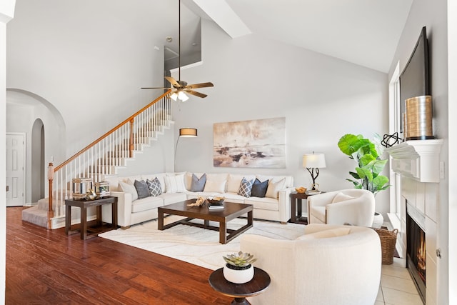 living room featuring a warm lit fireplace, stairway, arched walkways, light wood finished floors, and ceiling fan