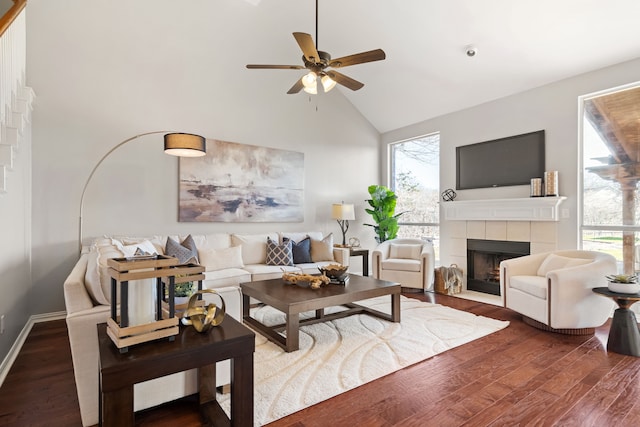 living area with high vaulted ceiling, a tiled fireplace, wood finished floors, baseboards, and ceiling fan