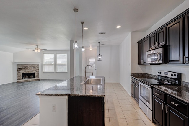 kitchen with a sink, ceiling fan, dark stone counters, stainless steel appliances, and a kitchen island with sink