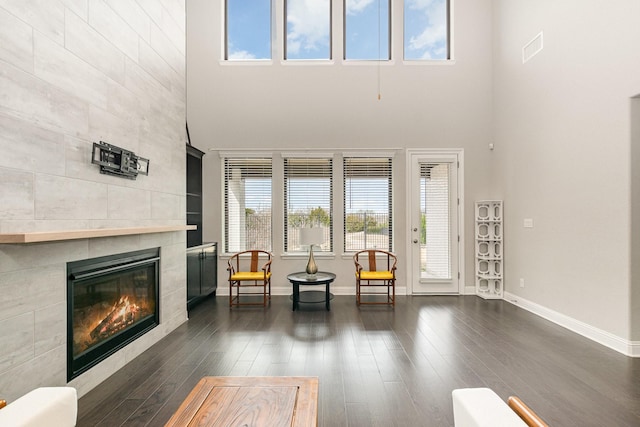 living area featuring a tiled fireplace, a high ceiling, baseboards, and dark wood-style flooring