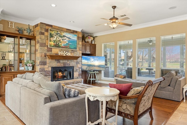 living area with ceiling fan, a wealth of natural light, crown molding, and a stone fireplace
