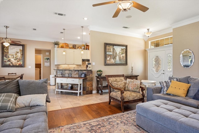 living room with light wood-type flooring, visible vents, ceiling fan, and crown molding
