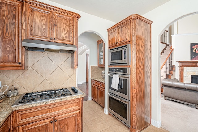 kitchen with tasteful backsplash, under cabinet range hood, brown cabinets, appliances with stainless steel finishes, and arched walkways