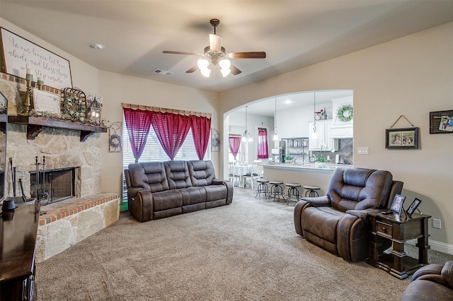 carpeted living room featuring visible vents, baseboards, ceiling fan, a stone fireplace, and arched walkways