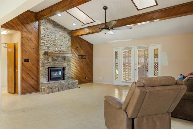 living area with a ceiling fan, beam ceiling, a skylight, a stone fireplace, and wood walls