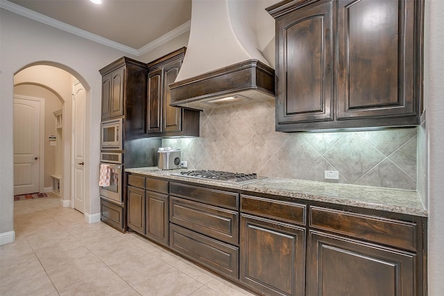 kitchen with custom range hood, light stone counters, stainless steel appliances, arched walkways, and dark brown cabinets