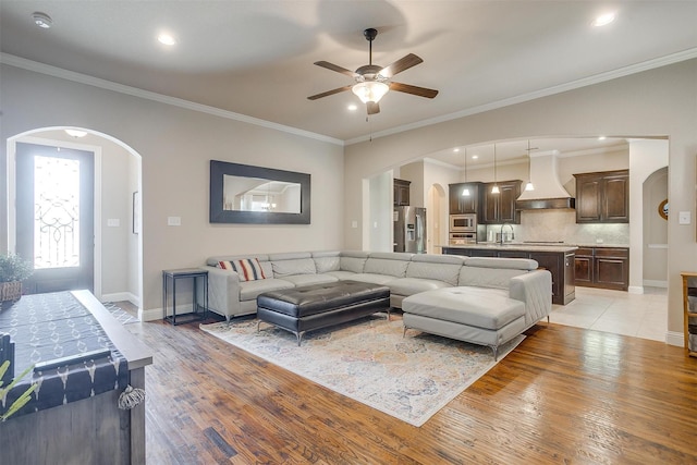 living room with light wood-style flooring, ornamental molding, arched walkways, baseboards, and ceiling fan