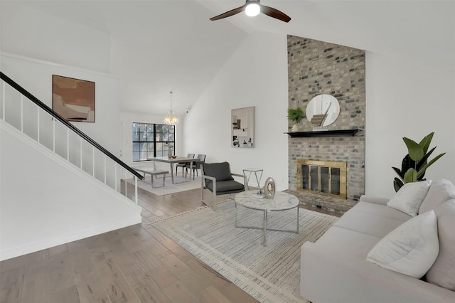 living room with stairway, wood finished floors, high vaulted ceiling, a brick fireplace, and ceiling fan with notable chandelier