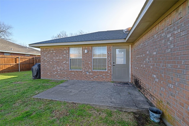 doorway to property featuring fence, a yard, a shingled roof, a patio area, and brick siding