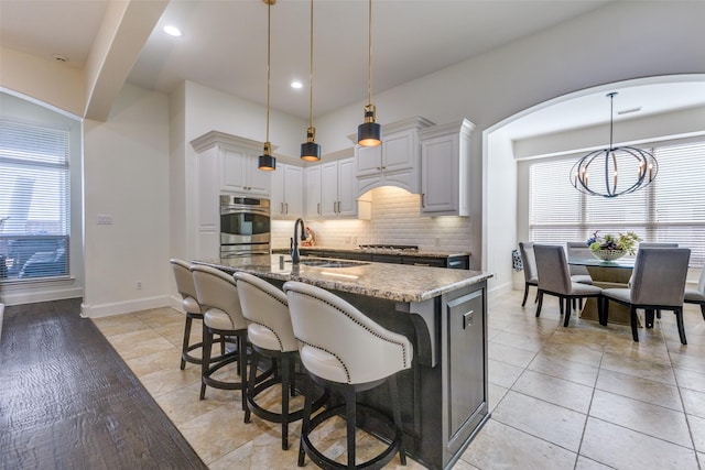 kitchen with an island with sink, decorative backsplash, arched walkways, a notable chandelier, and a sink