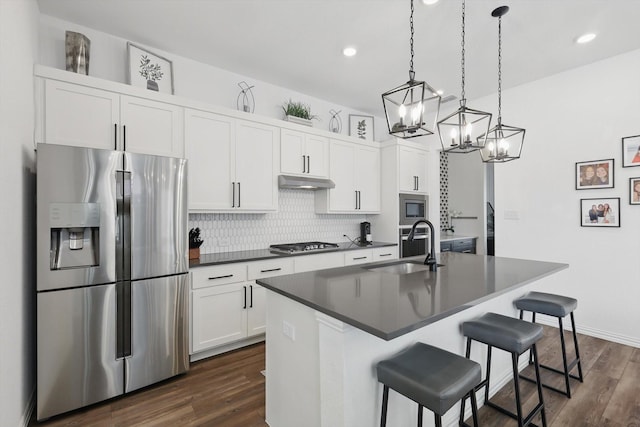 kitchen with a kitchen island with sink, under cabinet range hood, a sink, dark countertops, and stainless steel appliances