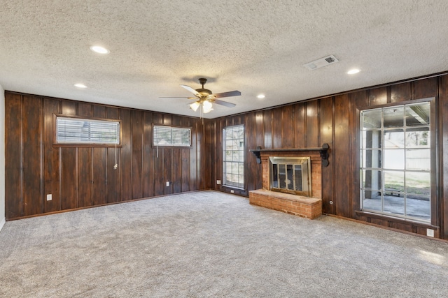 unfurnished living room with visible vents, a ceiling fan, wooden walls, carpet flooring, and a brick fireplace