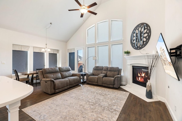 living room with high vaulted ceiling, ceiling fan with notable chandelier, a glass covered fireplace, dark wood finished floors, and baseboards