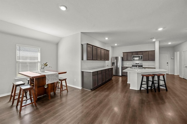 kitchen featuring a kitchen island with sink, dark brown cabinets, light countertops, stainless steel appliances, and dark wood-style flooring