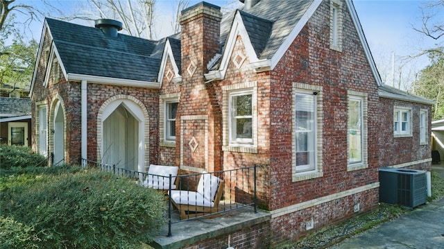 tudor house with brick siding, central air condition unit, a chimney, and a shingled roof