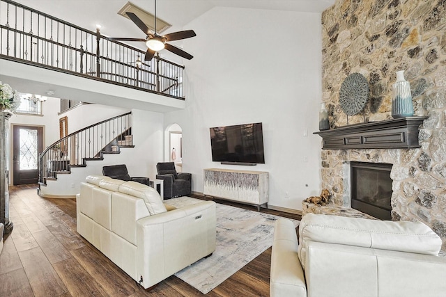 living area with a ceiling fan, arched walkways, dark wood-style flooring, stairs, and a stone fireplace