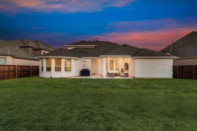 back of house at dusk with a shingled roof, a lawn, a fenced backyard, and a patio area