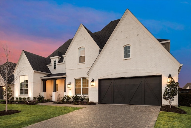 french country home with brick siding, decorative driveway, a yard, and roof with shingles