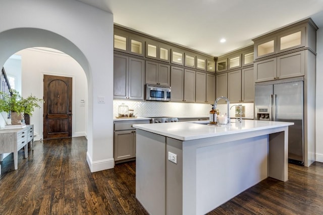 kitchen featuring a sink, backsplash, arched walkways, stainless steel appliances, and a kitchen island with sink