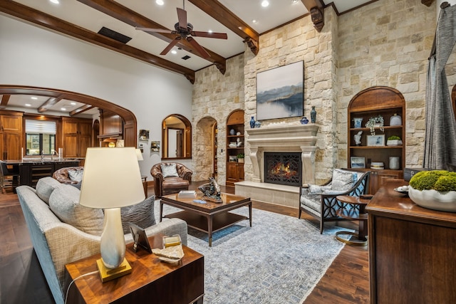 living area featuring a ceiling fan, dark wood finished floors, beam ceiling, arched walkways, and a stone fireplace