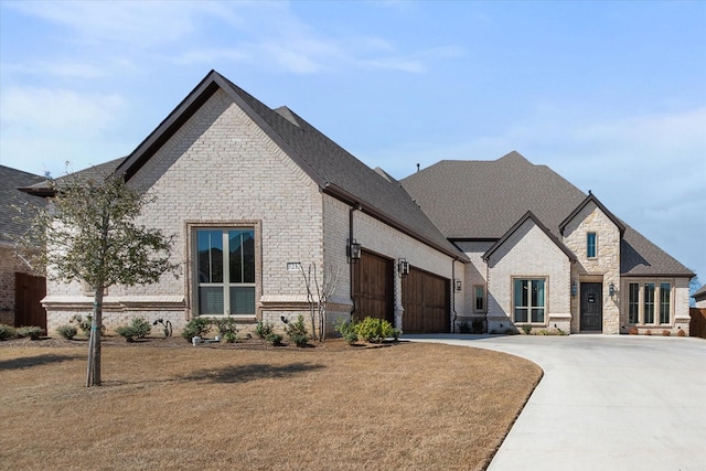 french country inspired facade featuring a front lawn, driveway, stone siding, an attached garage, and brick siding