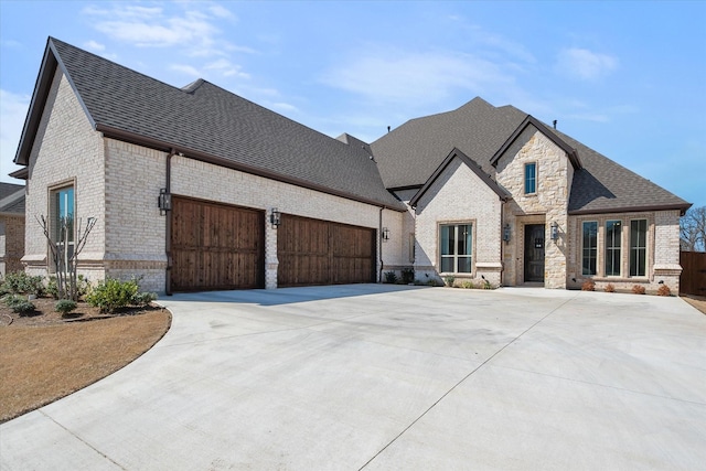 french provincial home with roof with shingles, an attached garage, concrete driveway, stone siding, and brick siding