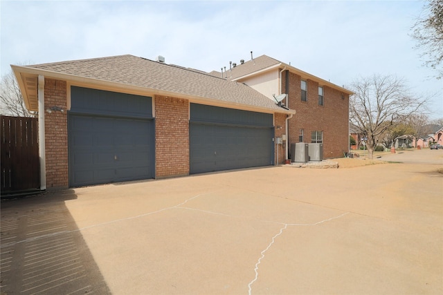 view of side of home featuring driveway, cooling unit, a shingled roof, a garage, and brick siding