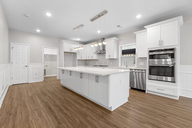 kitchen with a sink, a kitchen island, appliances with stainless steel finishes, white cabinets, and wall chimney range hood