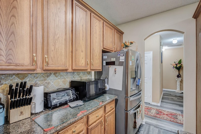 kitchen featuring arched walkways, stainless steel fridge, brown cabinetry, black microwave, and decorative backsplash